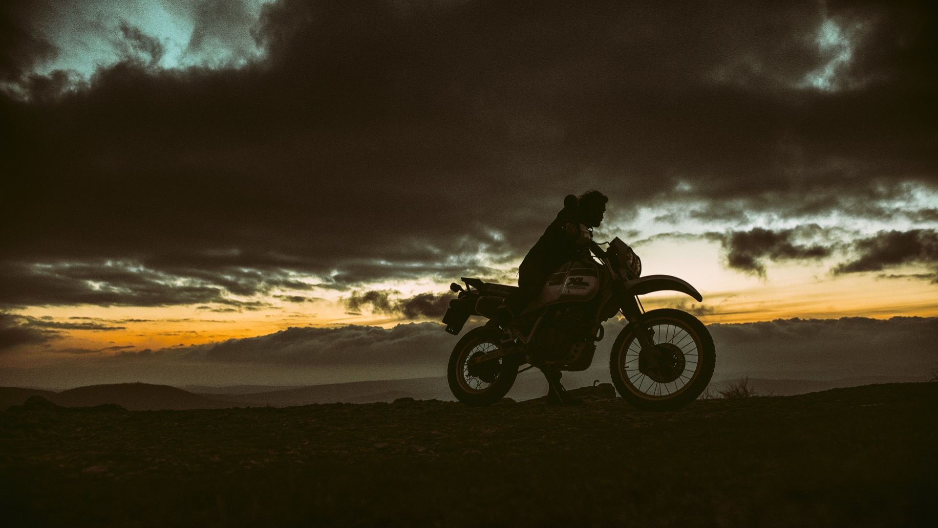 A rider sits on a vintage dual-sport motorcycle, silhouetted against a stormy sunset sky.