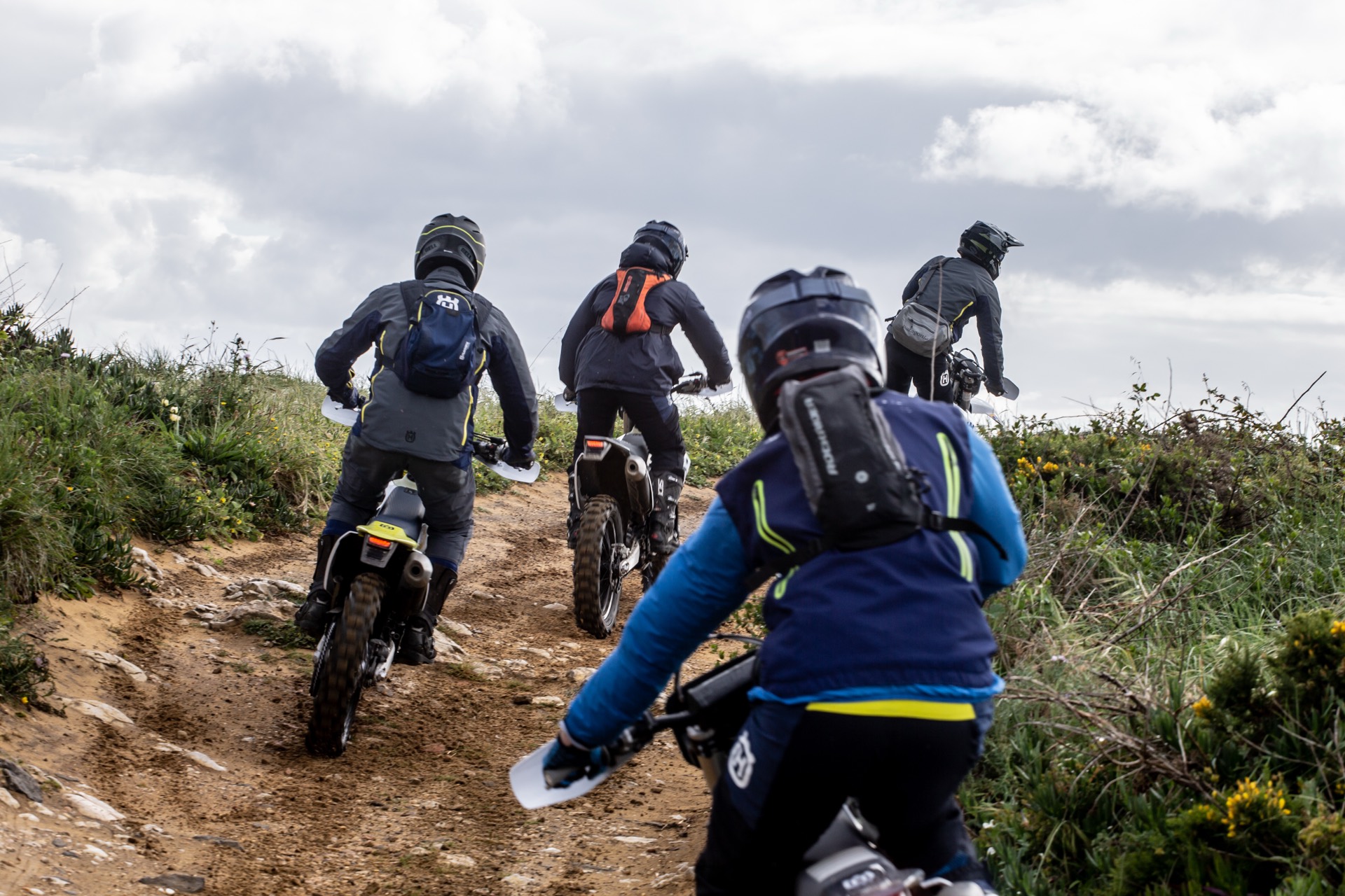 Riders head out together along a coastal dirt track, seen from behind.