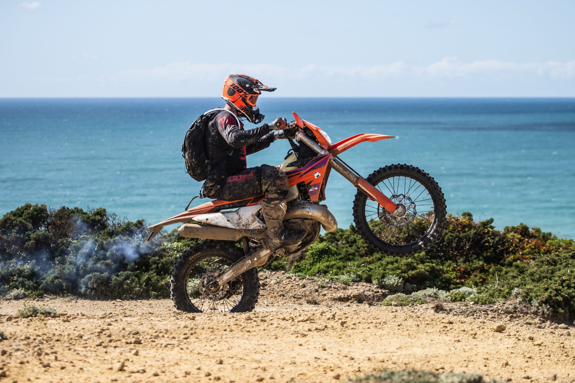 A rider on an adventure bike crosses a sandy trail beside the sea.