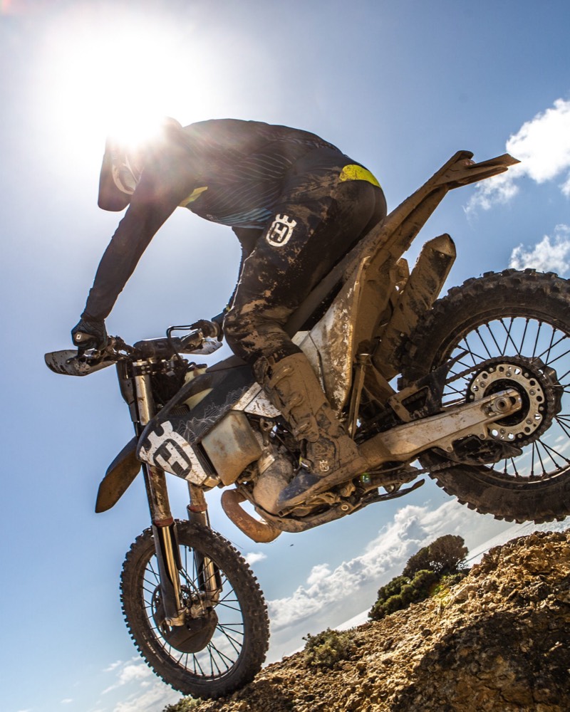 An off-road rider launches over a sandy ridge against a bright sky.